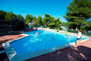Schwimmbad eines Campingplatzes mit Kindern im Wasser, bunten Spielrutschen und Liegefläche, umgeben von Bäumen und Blumenbeeten.