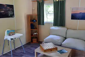 Wohnbereich im Camping-Unterkunft: graues Sofa, Fenster mit grünen Vorhängen, Kinderhochstuhl und Holzregal mit Spielzeug.