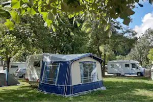 Wohnwagen und Vorzelt auf dem Campingplatz unter großen Bäumen, im Hintergrund weitere Camper und ein Parkplatz.