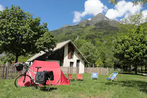 Rotes Campingzelt vor einem kleinen Gebäude, umgeben von Bäumen, mit Blick auf einen hohen Berg im Hintergrund.