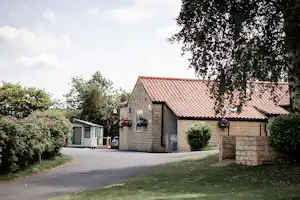 Campingplatz-Gebäude mit Ziegeldach, Einfahrt und Rasenfläche, daneben ein Wohnwagen und Hecken im Garten.