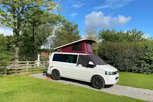 Weißes Wohnmobil auf einem Kiesweg neben einer grünen Campingwiese, umgeben von Bäumen und Hecken.