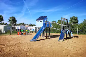 Spielplatz am Campingplatz mit blauer Rutsche, Klettergerüst und Sandboden, im Hintergrund weiße Wohnwagen und Bäume.