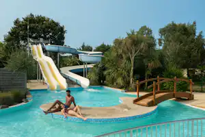 Kinderpool mit gelber Rutsche, Palmen und Holzgeländer am Campingplatz, daneben Liegefläche und Wasserbecken.