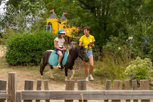 Familie auf einem Pferd im Grünen, Kinder und Erwachsener reiten auf dem Campingplatz neben Hecken und Zaunpfosten.