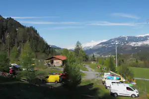 Campingplatz im Tal: Wohnwagen und Autos am Weg, kleines Gebäude mit rotem Dach, Bäume und Bergpanorama im Hintergrund.