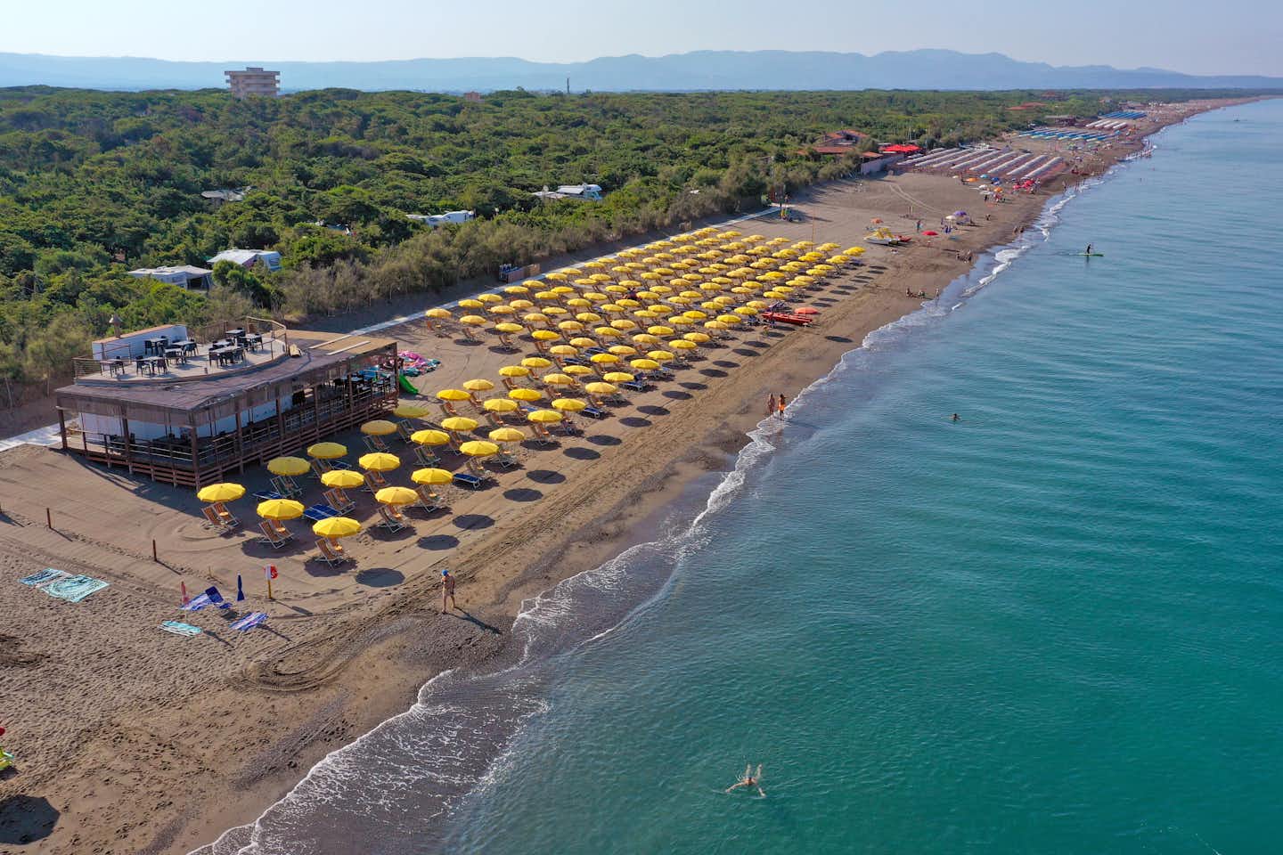 Camping Village Casa di Caccia - Vogelperspektive auf den Strand mit Liegestühlen, Sonnenschirmen und Strandbar und teilweise erkennbare Standplätze zwischen den Bäumen