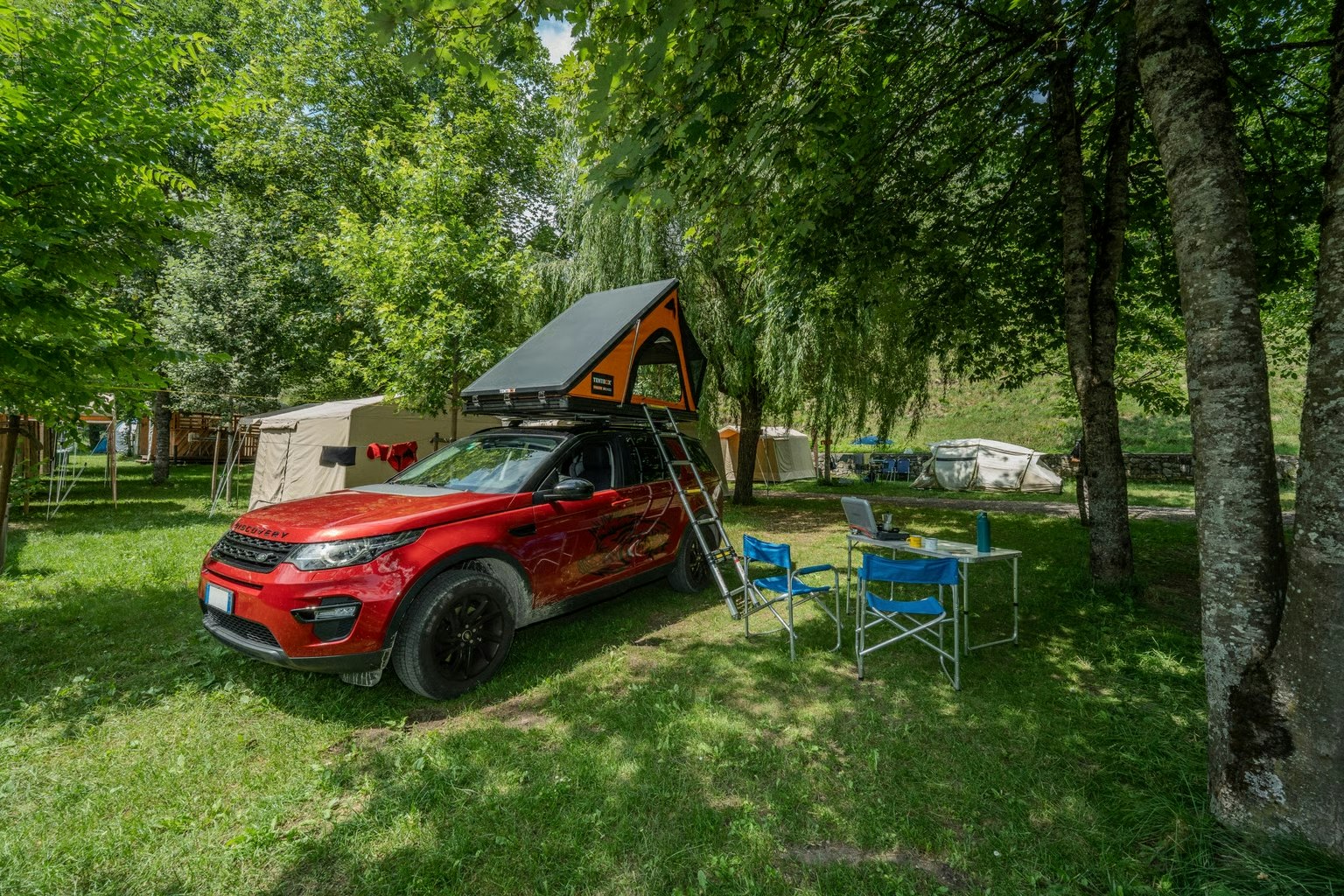 Roter Camper mit Vorzelt am Stellplatz im Grünen, daneben blaue Campingstühle und Zelt, umgeben von Bäumen.