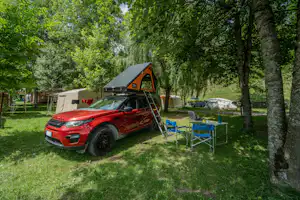 Roter Camper mit Vorzelt am Stellplatz im Grünen, daneben blaue Campingstühle und Zelt, umgeben von Bäumen.