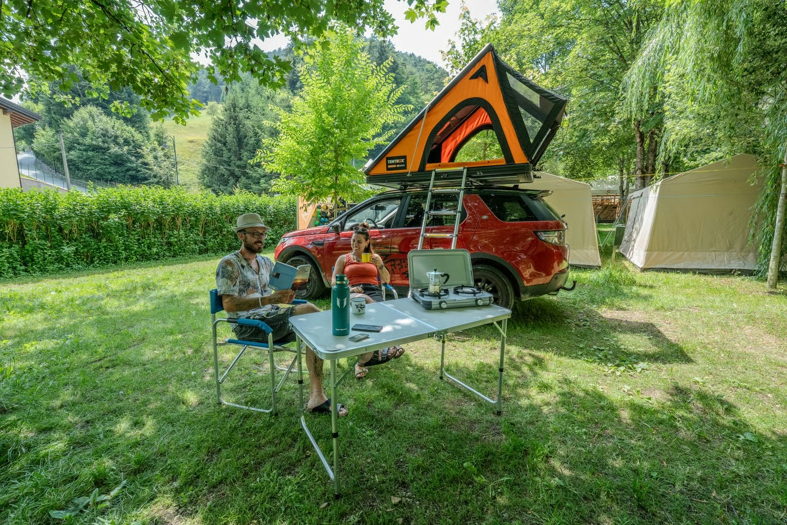 Campingplatz mit Zelt und Wohnmobil, davor Tisch und Stühle im Grünen, daneben ein Mann am Campingstuhl.