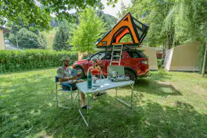 Campingplatz mit Zelt und Wohnmobil, davor Tisch und Stühle im Grünen, daneben ein Mann am Campingstuhl.