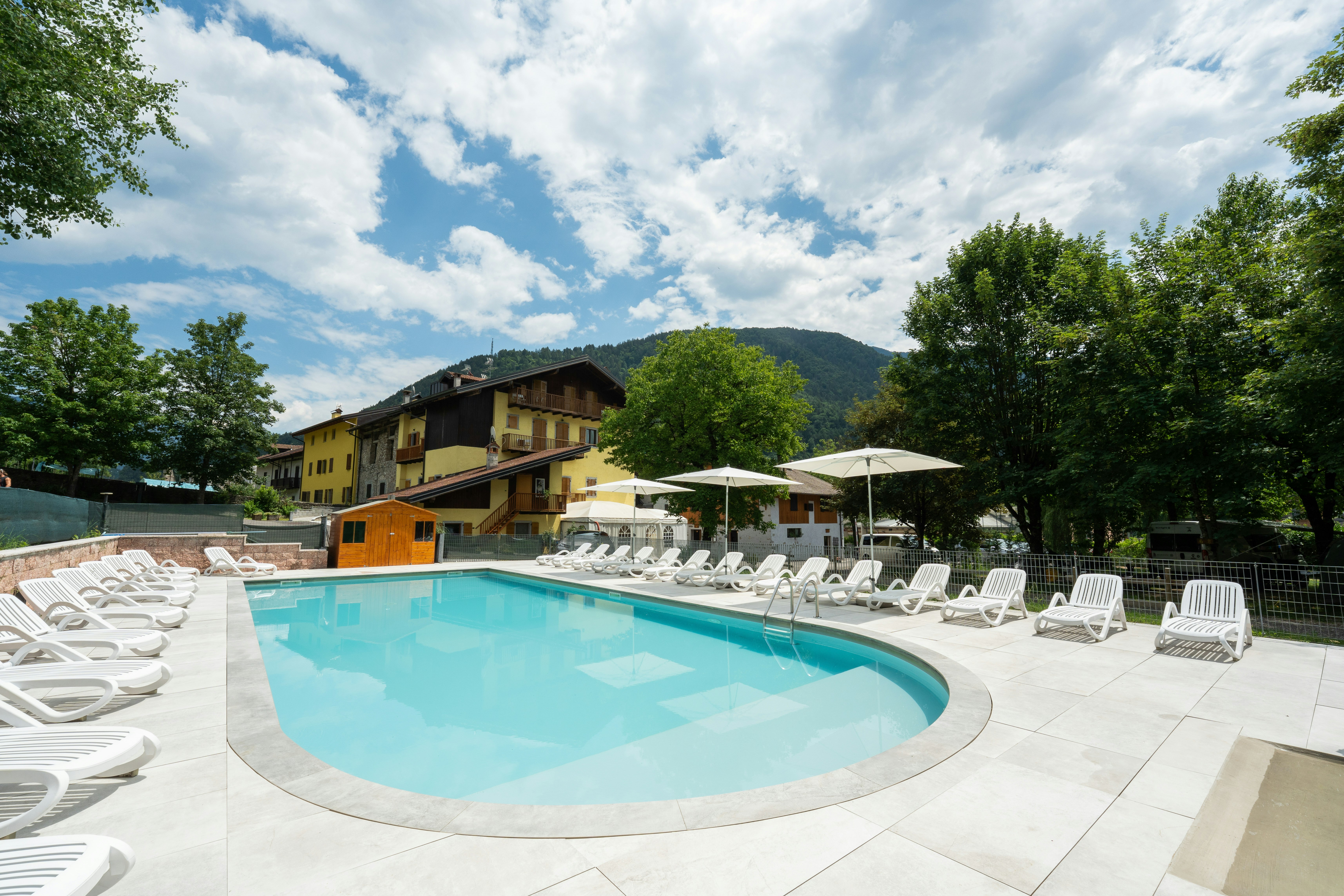 Schwimmbad mit rundem Becken und Liegestühlen auf der Terrasse, umgeben von Bäumen und einem mehrstöckigen Gebäude im Hintergrund.