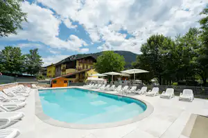 Schwimmbad mit rundem Becken und Liegestühlen auf der Terrasse, umgeben von Bäumen und einem mehrstöckigen Gebäude im Hintergrund.