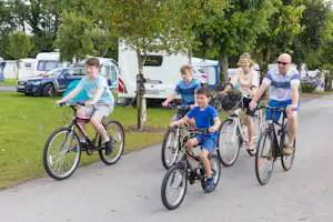 Camping Caseys - radfahrende Familie auf dem Campingplatz mit Wohnwagen im Hintergrund