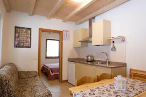 Kleine Küchenzeile mit Spüle und Herd, daneben Sofa und Esstisch im hellen Wohnbereich mit Holzbalkendecke.
