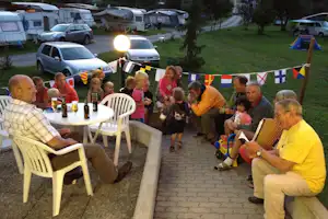 Campingplatz-Abend: Tisch mit Getränken und Stühlen, im Hintergrund Wohnwagen und Autos auf dem Stellplatz.