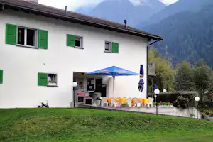Campinghaus mit grünen Fensterläden, Terrasse mit Sonnenschirm und Stühlen, im Hintergrund bewaldete Berge.