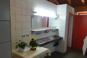Waschraum mit langen Waschbecken auf Fliesenwand, Wasserhähnen, Tisch mit Blumen und Blick in den Durchgang.