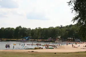 Badestrand am See mit vielen Schwimmern, gelben und grünen Luftmatratzen, Steg und Bäumen im Hintergrund.