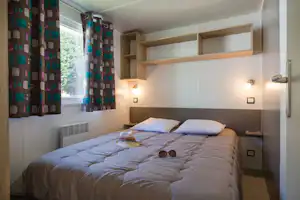 Doppelbett im Campingzimmer mit Holzregal über dem Kopfteil, Vorhängen am Fenster und warmen Lampen an der Wand.