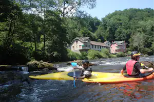 Kajaks auf einem Fluss mit Stromschnellen, im Hintergrund Gebäude und viel Wald, eine Person paddelt im Vordergrund.