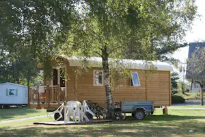 Holzbungalow mit weißem Dach, Veranda und Treppe; davor weiße Gartenstühle, Fahrradanhänger und Bäume im Campingbereich.