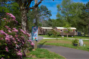 Mobilheim mit Vorzelt und Fenstern, neben einem Wohnwagen auf dem Campingplatz, umgeben von Bäumen und Rasen.