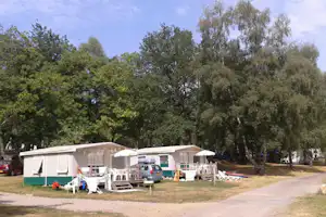 Campingstellplätze mit Wohnwagen und Vorzelten, Tischen und Stühlen vor grünen Bäumen entlang eines Weges.