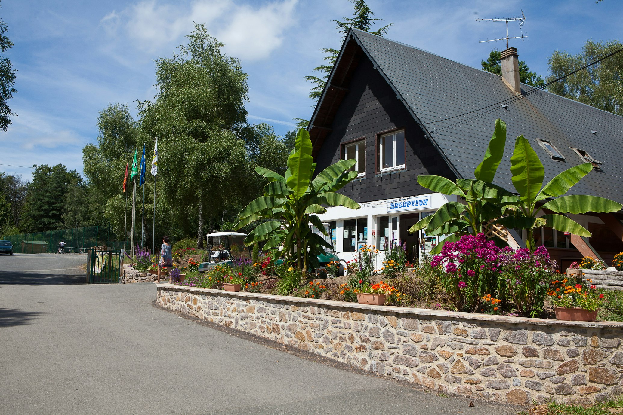 Rezeption eines Campingplatzes mit Holzhaus, Blumenbeeten und Palmen vor dem Eingang, im Hintergrund Bäume und Fahnen.