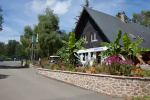 Rezeption eines Campingplatzes mit Holzhaus, Blumenbeeten und Palmen vor dem Eingang, im Hintergrund Bäume und Fahnen.