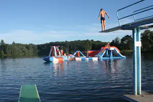 Wasserrutsche und aufblasbares Wasser-Spielgerät im See, daneben ein Sprungturm mit Person auf der Plattform.