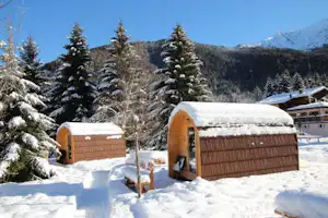 Holz-Glampinghütten im Schnee mit verschneiten Bäumen und Blick auf die Berge, im Hintergrund ein Gebäude am Campingplatz.