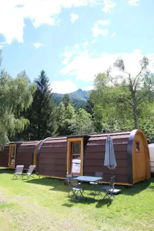 Buntes Zelt mit Holzfront und Sonnenschirm auf einem Campingplatz, daneben Liegestühle auf grünem Rasen und Bäume im Hintergrund.
