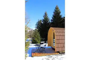 Holzfass-Sauna auf verschneitem Campingplatz mit Sitzbank und Tisch, umgeben von Nadelbäumen und blauem Himmel.
