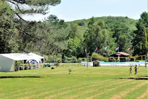 Großer Campingplatz mit Rasen, Zelt und Schwimmbecken, umgeben von Bäumen und Hügeln im Hintergrund.