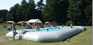 Aufblasbares Planschbecken auf einer Wiese neben einem Zelt und Sonnenschirmen, im Hintergrund Bäume und ein Poolbereich.