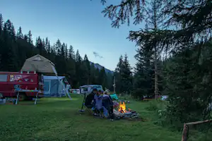 Campingplatz mit rotem Camper, aufgestelltem Zelt und Lagerfeuer am Tisch, umgeben von Tannen und Bergen im Hintergrund.