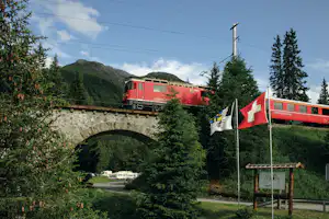 Roter Zug fährt über eine Steinbrücke, im Vordergrund Campingplatz mit Wohnmobilen, Bäumen und Schildern.