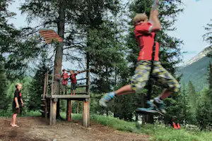 Kinder auf einer Holzplattform am Spielplatz, eine Person rutscht auf der Rutsche durch den Wald.