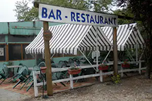 Schild „BAR - RESTAURANT“ über einer Terrasse mit gestreifter Markise, grünen Plastikstühlen und Pflanzkübeln.