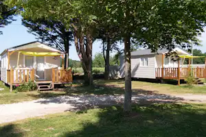 Zwei Mobilheime mit Holzterrassen, Markisen und Sitzbank, umgeben von Bäumen und Rasen auf dem Campingplatz.