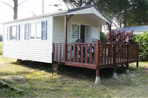 Mobilheim mit Holzveranda, Stühlen am Eingang und blühendem Strauch neben dem Haus im Grünen.