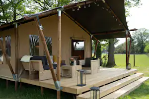 Holzterrasse am Zelt mit überdachtem Vorbau, Sitzbank und Kissen, daneben Holzboxen und Blick in den Garten mit Bäumen.