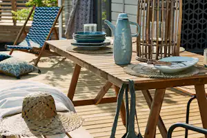 Holzterrasse mit Tisch und Geschirr, daneben Deko-Laterne, Kaffeekanne und Stuhl, im Hintergrund Liegestuhl und Vorhang am Gebäude.