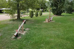 Grüne Campinganlage mit Kiesweg, Bäumen und Hecken, im Vordergrund ein Holzpfosten mit Absperrband.