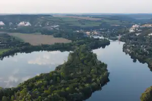 Breiter Fluss mit ruhigem Wasser, umgeben von Wäldern, Feldern und einem Dorf in der Ferne.