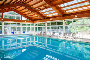 Innenpool mit blau gefliester Beckenwand, Liegen und großen Fenstern unter Holzdecke im Campinghaus.
