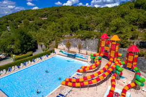 Großes Camping-Schwimmbecken mit bunten Rutschen und Spielbereich, Liegen am Rand und Waldhügeln im Hintergrund.