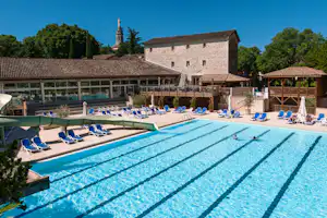 Großer Camping-Pool mit blauen Liegen, Sonnenschirmen und Blick auf Gebäude und Bäume im Hintergrund.