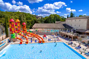 Großes Camping-Schwimmbecken mit Liegen, Sonnenschirmen und buntem Wasser-Spielbereich, umgeben von Gebäuden und Bäumen.